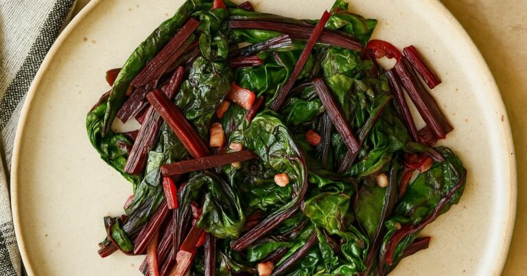 A plate of sautéed Swiss chard featuring vibrant green leaves and reddish stems, garnished with small pieces of bacon.